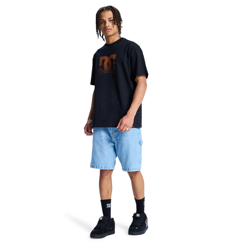 A young man with braided hair stands against a white background, wearing a black Reebok DC Star Oxidized Hss t-shirt, light blue denim shorts, black socks, and black sneakers.