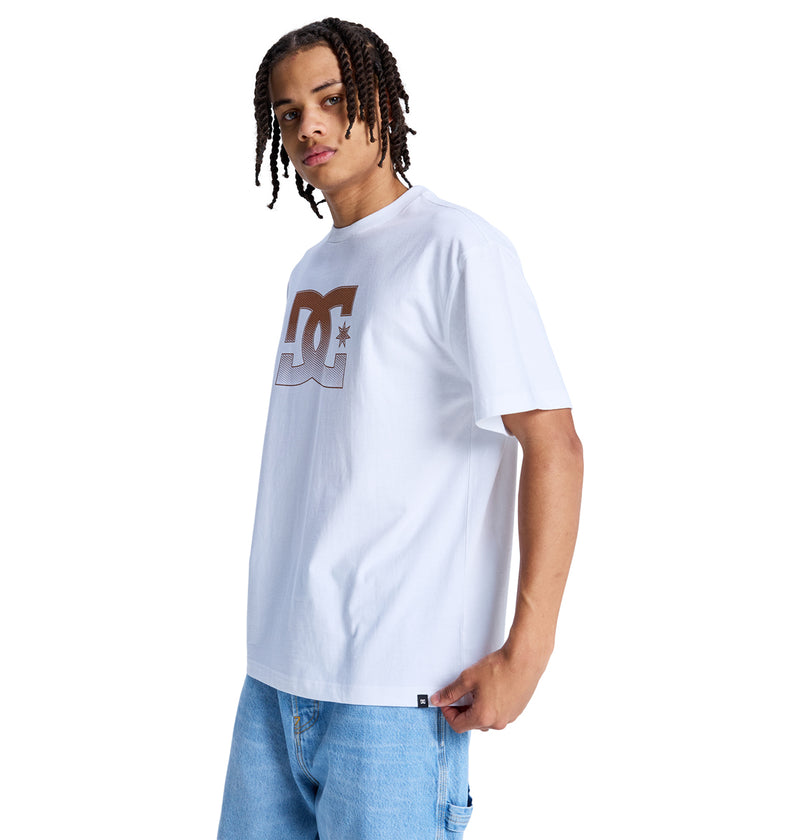 A person wearing the Reebok DC Star Oxidized Hss white t-shirt with a large brown DC logo on the chest and light blue jeans stands against a white background, facing slightly to the side.