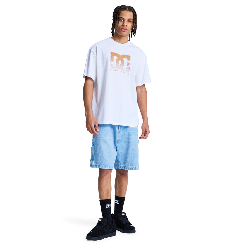 A young man with braids stands against a white background wearing a white Reebok DC Star Oxidized Hss t-shirt, light blue denim shorts, black socks, and black sneakers.