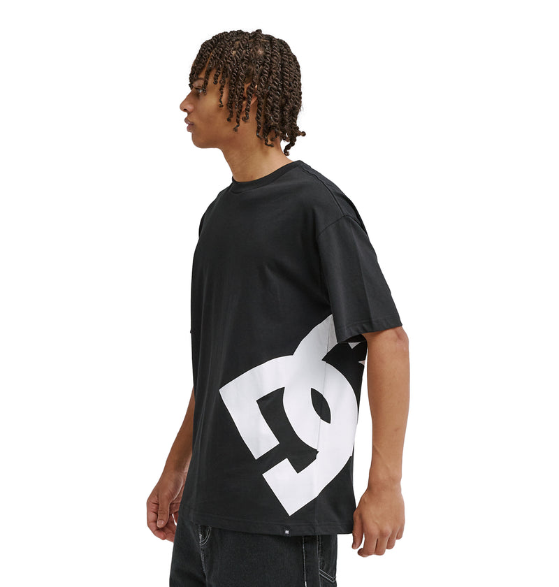 A person with short, curly hair stands sideways against a plain white background, wearing the Reebok DC Lanai Hss—a loose black T-shirt featuring a large white graphic logo on the lower side—and black jeans.