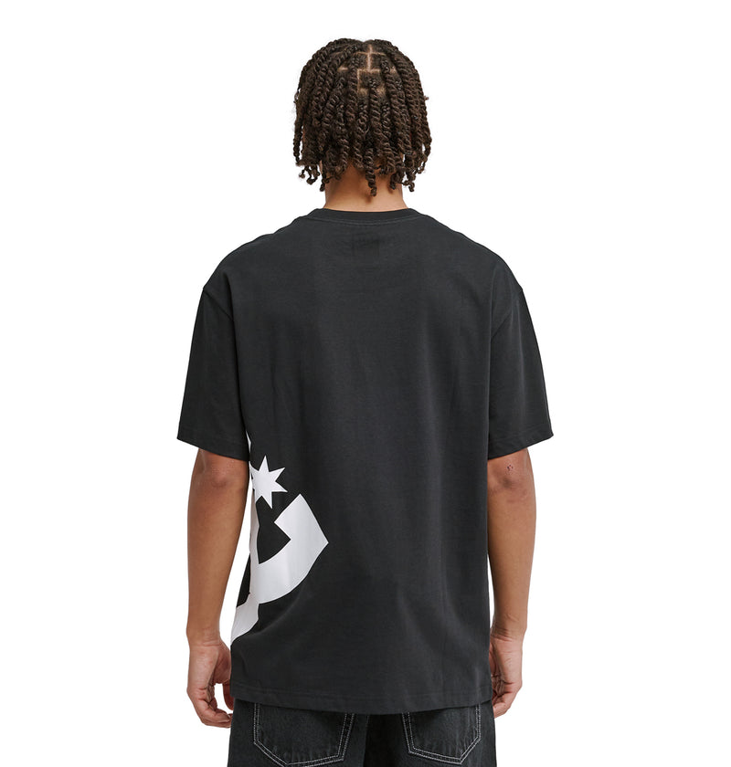 A person with short, twisted braids is wearing the Reebok DC Lanai Hss, a loose black T-shirt featuring a bold white graphic on the lower back, standing against a plain white background and facing away from the camera.