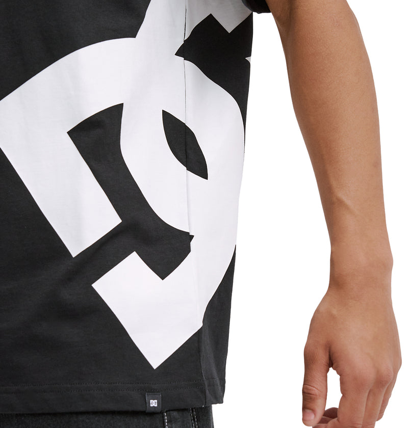 A person shows the torso and right arm while wearing the Reebok DC Lanai Hss, a black T-shirt with a large, bold white graphic design; their hand is relaxed at their side.