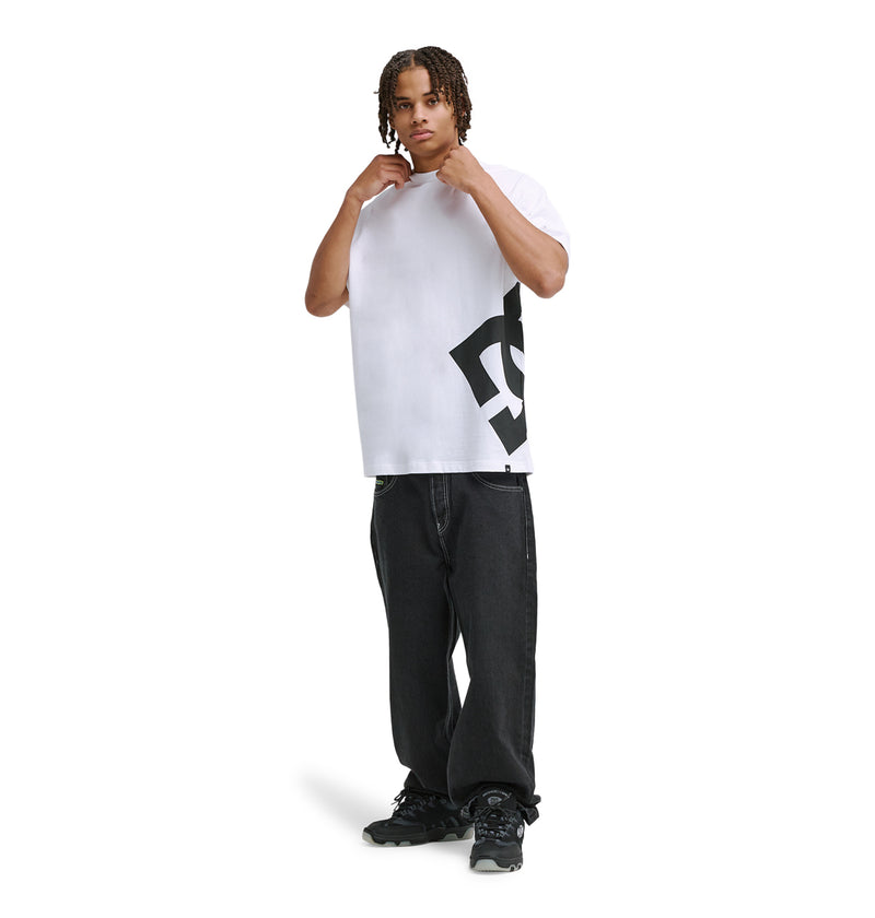 A person with short braids adjusts their collar while wearing the Reebok DC Lanai Hss—a white tee with a bold black graphic—paired with loose black jeans and black sneakers, all set against a white background.