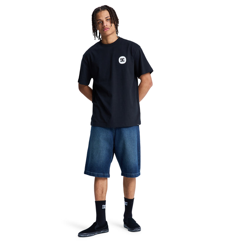 A person with short, braided hair stands facing forward against a white background, wearing a black Shape Or Destroy Hss t-shirt by Reebok, knee-length blue denim shorts, black socks, and black slip-on shoes.