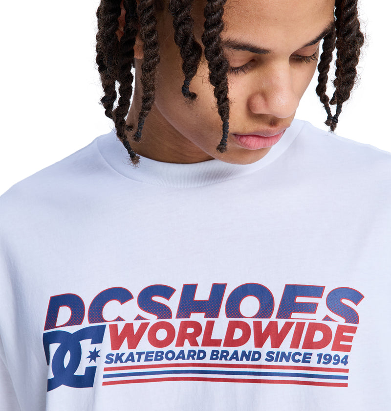 A person with braided hair looks down while wearing a white Queltra Lhss T-shirt by Reebok, featuring bold blue and red text on the front.
