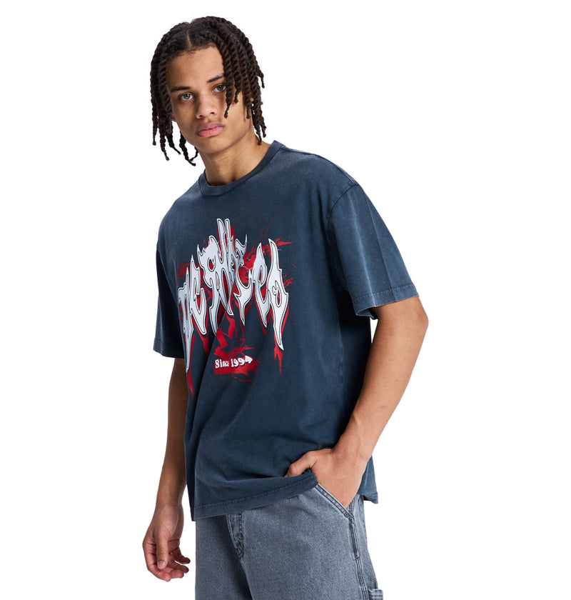 A young person with braided hair wears a loose-fitting, faded black DC Death Machine Hss graphic T-shirt by Reebok and light blue jeans. They stand with one hand in their pocket, facing the camera.