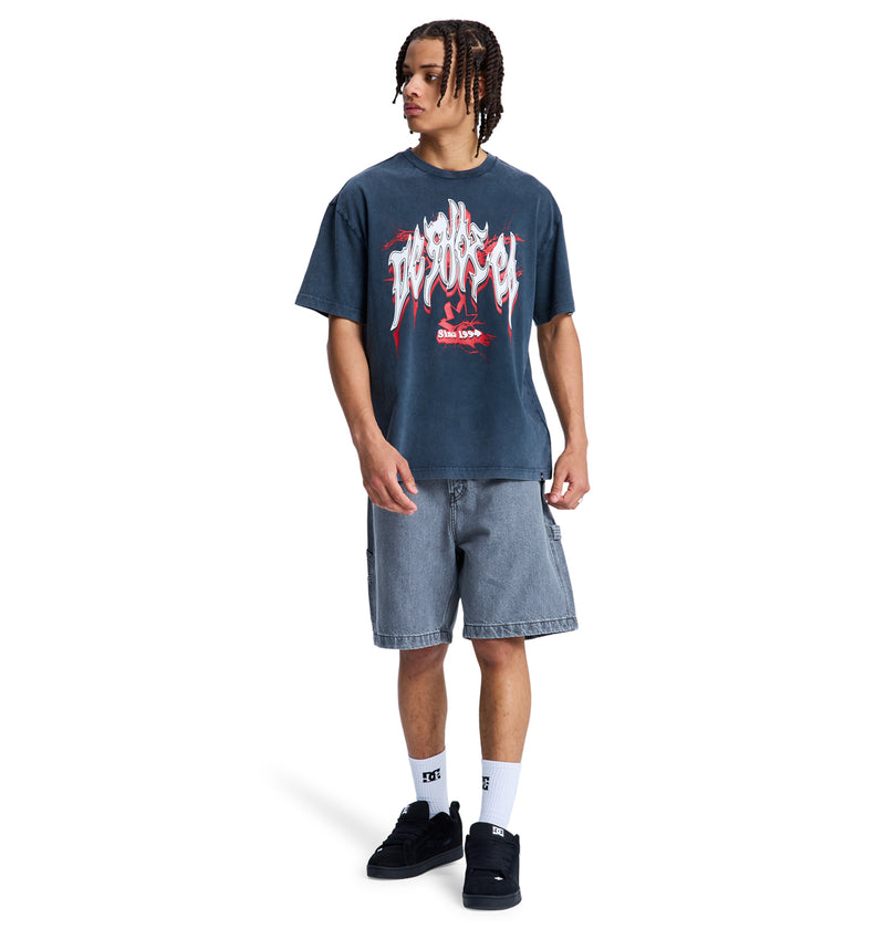 A person with braided hair wears the Reebok DC Death Machine Hss graphic T-shirt, loose gray denim shorts, white socks, and black sneakers, standing against a white background.