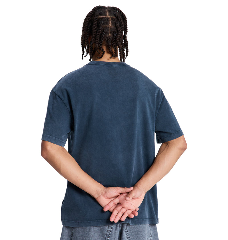 A person with braided hair, seen from behind, wears a loose-fitting, short-sleeved DC Death Machine Hss t-shirt by Reebok in dark blue and blue jeans, with their hands clasped behind their back.