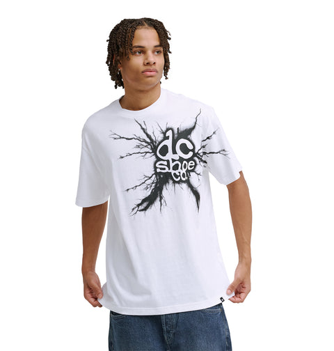 A young man with short dreadlocks wears the Reebok Strike Twice Hss loose white T-shirt with a black splatter-style logo on the chest, paired with loose blue jeans, standing against a plain white background.