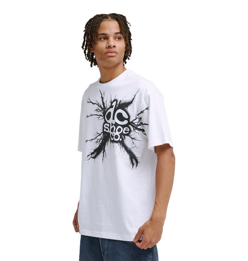 A young person with short dreadlocks wears a loose white Reebok Strike Twice Hss T-shirt with a bold black graphic, standing against a plain white background and looking to the side.