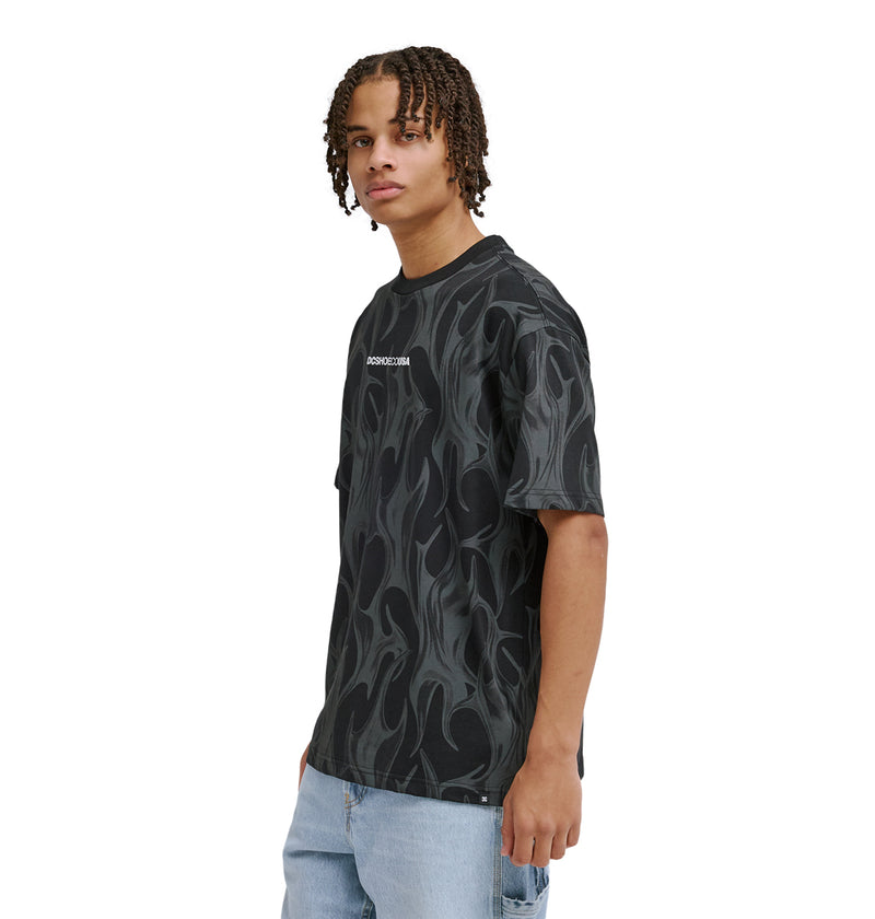 A young person with short dreadlocks wears the Reebok DC Liquid Fuego Hss black t-shirt with a gray flame pattern and light blue jeans, standing against a white background.