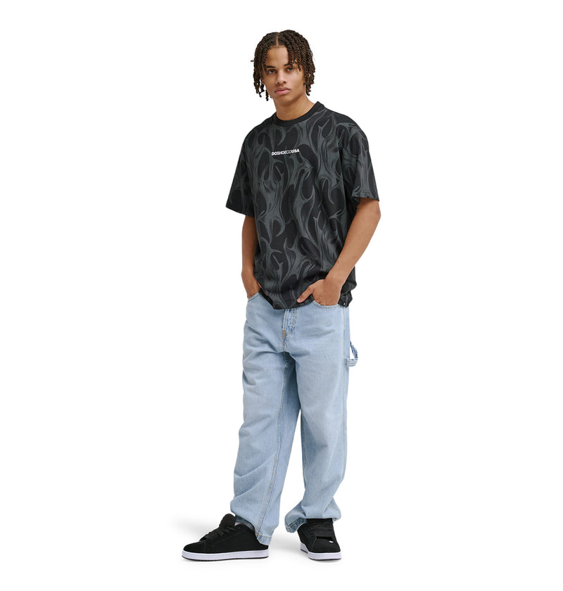 A young person with short, curly hair stands facing forward in a relaxed pose, wearing the Reebok DC Liquid Fuego Hss T-shirt with black flame patterns, light blue baggy jeans, and black sneakers. One hand is casually in their pocket.