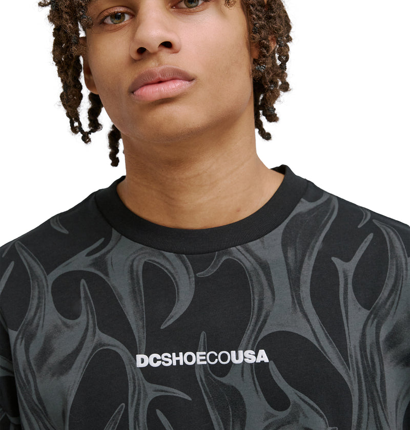 A young person with medium skin tone and short dreadlocks wears the Reebok DC Liquid Fuego Hss, a black shirt with a gray flame pattern and DCSHOECOUSA in white on the chest, set against a plain white background.