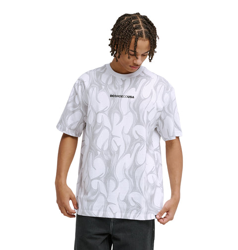 A person with braided hair looks down, wearing the Reebok DC Liquid Fuego Hss white T-shirt featuring a light gray abstract flame pattern and "DCSRBCO.CUBA" text on the chest, paired with black pants.