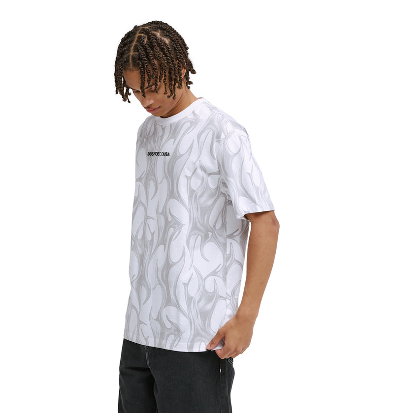 A person with braided hair wears the Reebok DC Liquid Fuego Hss white T-shirt featuring a subtle gray abstract flame pattern and small black chest text, paired with dark pants, looking down with one hand by their side.