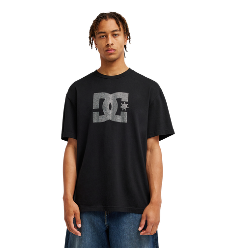 A person stands facing forward against a plain white background, wearing the Men’s DC Strass Star Tee by DC Shoes—a relaxed fit black graphic skate tee featuring a rhinestone DC star—paired with blue jeans.