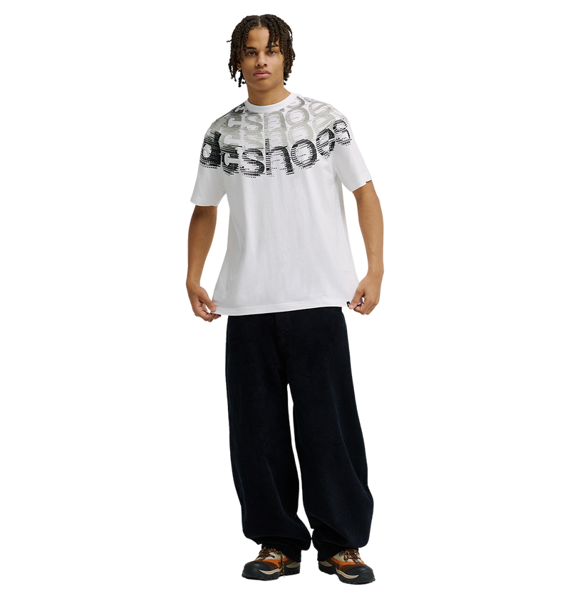 A person wearing the Men’s DC Omega 2 Tee from DC Shoes—a relaxed fit white graphic skate t-shirt with bold chest text—paired with loose black pants and brown sneakers, stands facing forward against a plain white background.