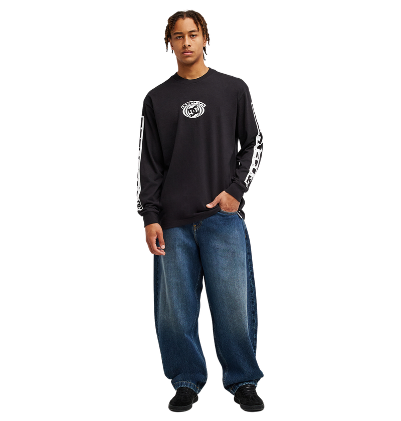 A person stands facing forward against a plain white background, wearing the Men's DC Echo Tee by DC Shoes—a black long-sleeve graphic tee featuring bold white DC-style graphics on the sleeves and chest—paired with loose blue jeans and black shoes, ready for casual skate sessions.
