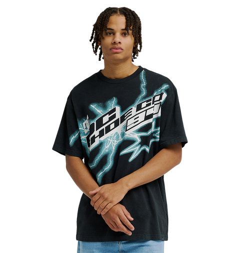 A man with short dreadlocks stands against a plain light background with his hands clasped in front, wearing the Men's DC Star Struck Tee by DC Shoes—a relaxed fit black skate shirt featuring bold, abstract white and teal graphics reminiscent of star struck artwork.