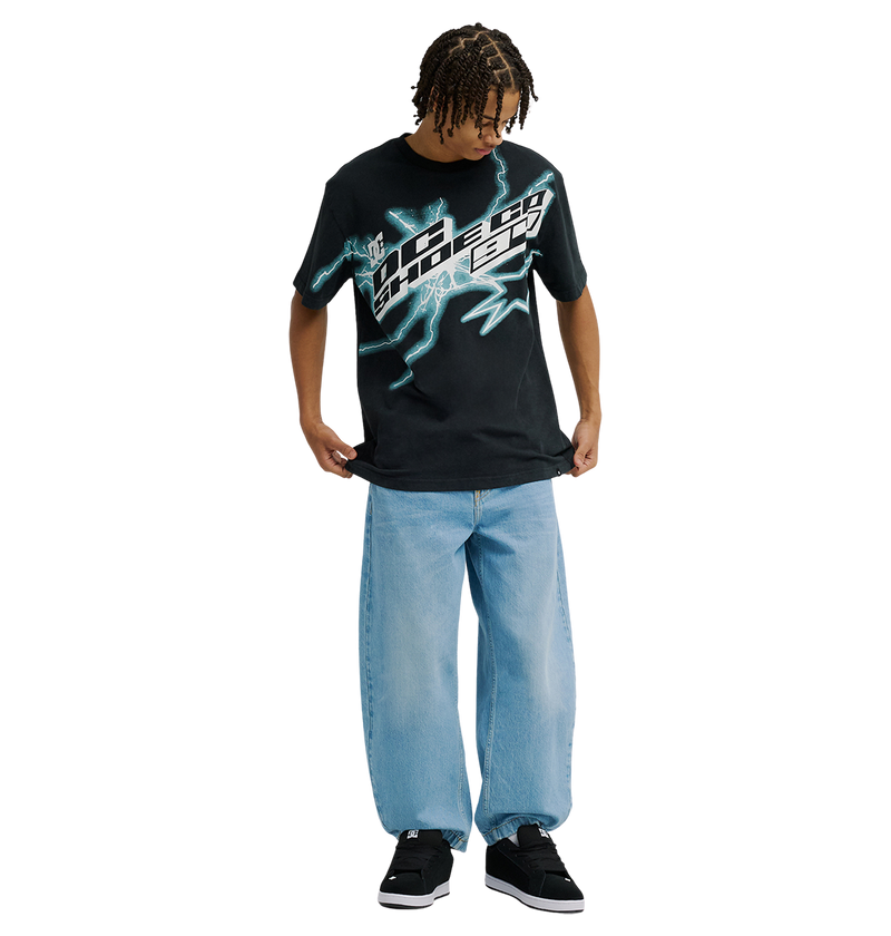 A person stands looking down, wearing the Men's DC Star Struck Tee by DC Shoes—a relaxed fit black graphic skate shirt featuring blue lightning designs—paired with light blue baggy jeans and black sneakers. The background is plain white.