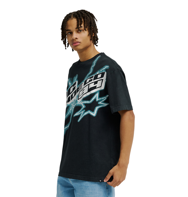 A man is seen wearing the Men's DC Star Struck Tee from DC Shoes—a black graphic skate T-shirt featuring bold blue lightning and starburst designs—while facing slightly to the side against a plain white background. The relaxed fit tee is styled with light blue jeans for an effortlessly cool look.