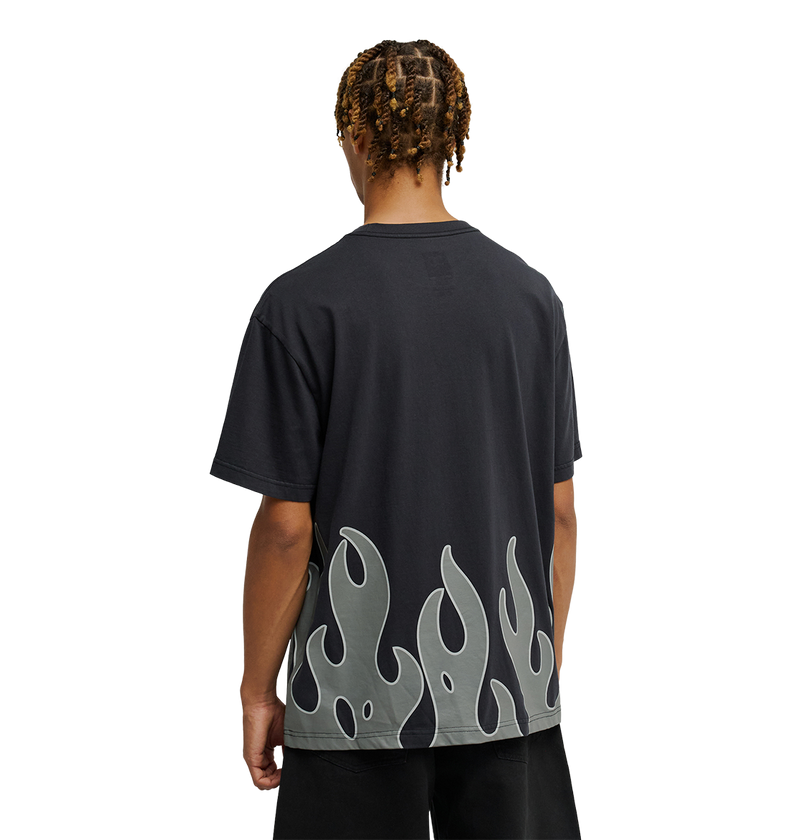 A person stands facing away, wearing the Men's DC Burning Diamonds Tee by DC Shoes—a black graphic skate shirt featuring a gray flame pattern along the bottom edge. The background is plain white.