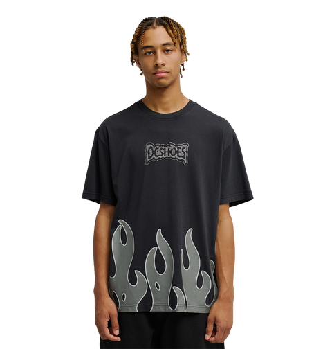 A young man with short braids wears the Men's DC Burning Diamonds Tee from DC Shoes—a black graphic skate t-shirt featuring the iconic Dogshoes logo on the chest and gray flame graphics at the hem, inspired by burning diamonds artwork. He stands out against a plain, light background, looking directly at the camera.