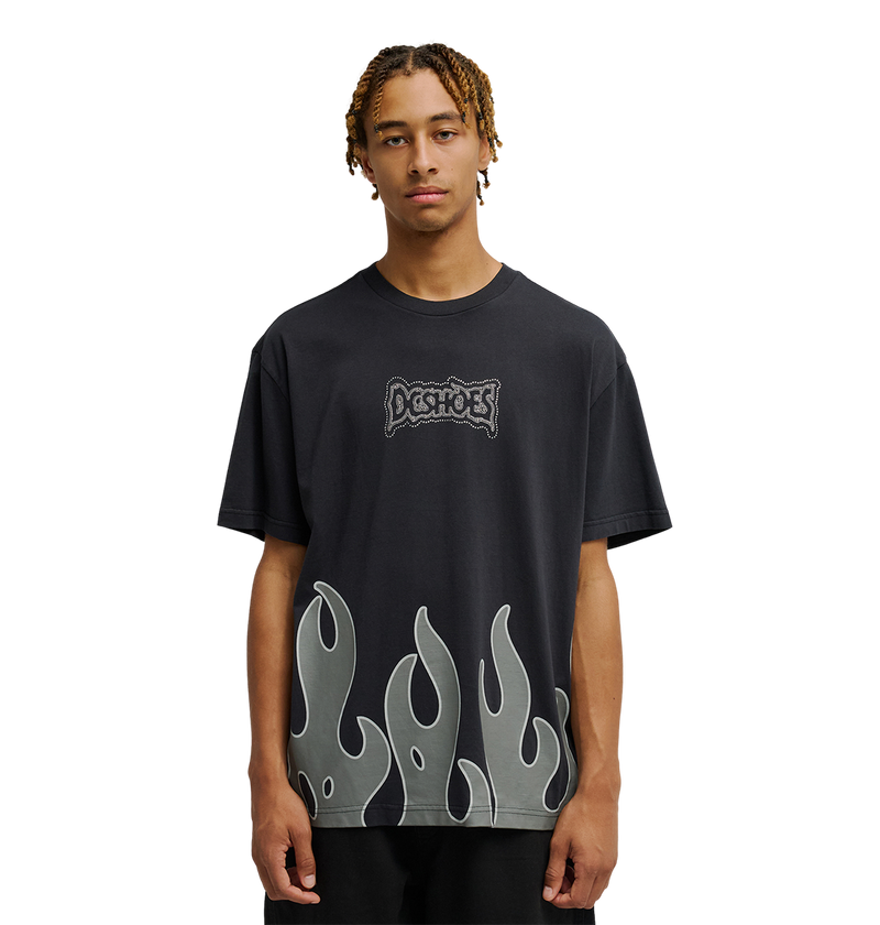 A young man with short braids wears the Men's DC Burning Diamonds Tee from DC Shoes—a black graphic skate t-shirt featuring the iconic Dogshoes logo on the chest and gray flame graphics at the hem, inspired by burning diamonds artwork. He stands out against a plain, light background, looking directly at the camera.