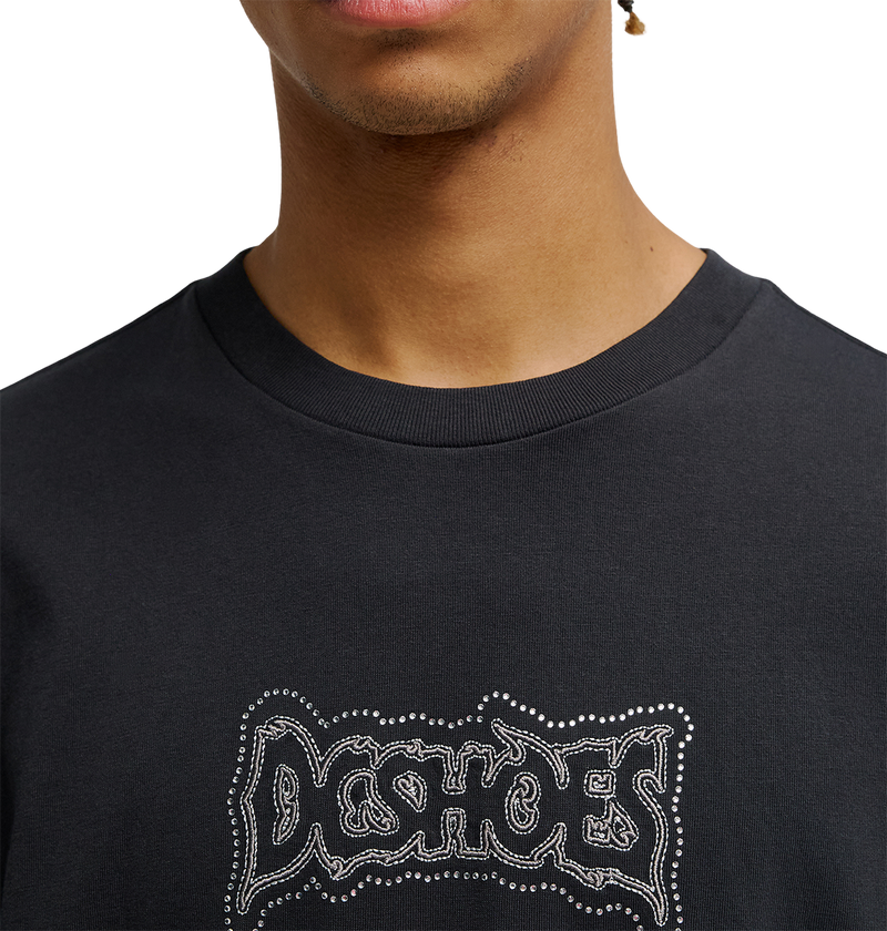 A person wearing the Men's DC Burning Diamonds Tee by DC Shoes, featuring a bold black graphic design with "DC SHOES" in a graffiti-style font accented by small white dots. Only the lower half of their face and upper torso are visible, emphasizing the streetwear aesthetic inspired by burning diamonds artwork.