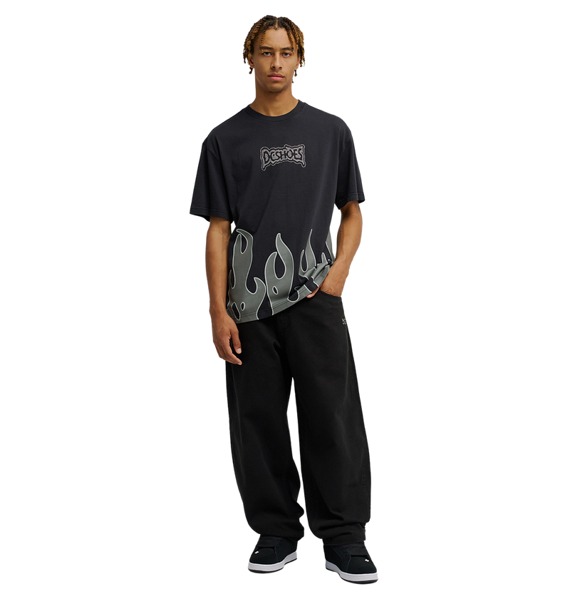 A person wearing the Men's DC Burning Diamonds Tee by DC Shoes, a black graphic skate shirt with a flame design along the bottom, and loose black pants stands facing forward against a plain white background.
