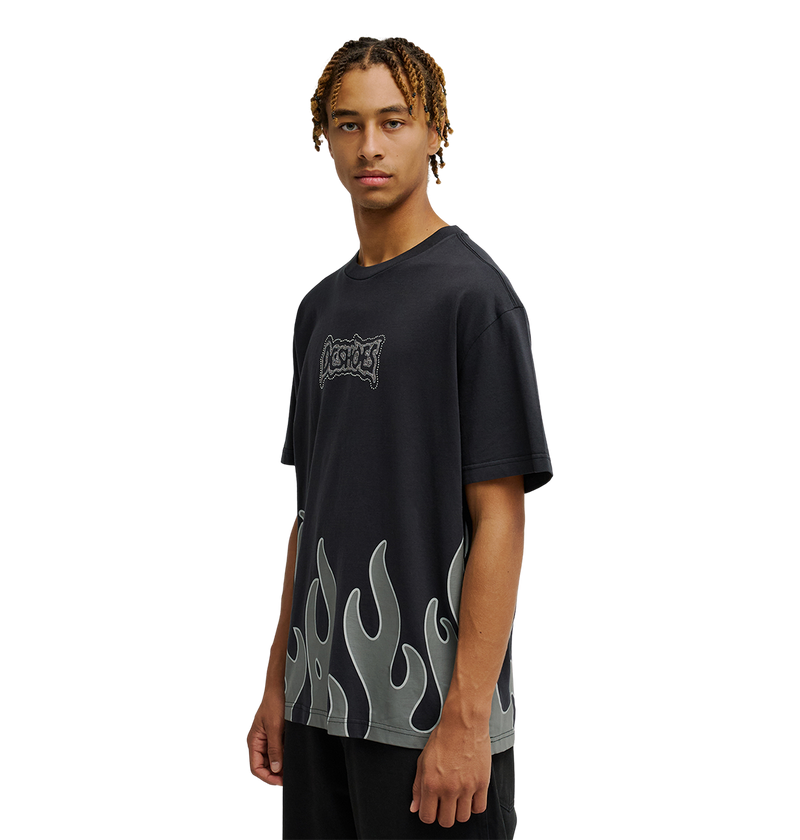 A person with short braided hair wears the Men's DC Burning Diamonds Tee by DC Shoes—a black graphic skate T-shirt with gray flame designs on the lower half and a small decorative logo on the chest—while standing against a plain white background.