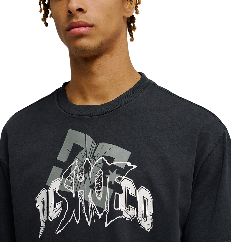 A person wearing the Men's DC Speratic Tee by DC Shoes—a black, relaxed-fit skate t-shirt featuring a bold, stylized gray and white "DC SHOCO" cracked-effect graphic. The heritage-inspired artwork stands out while only the lower half of the face is visible.