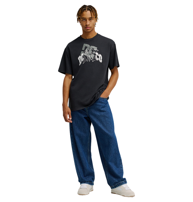 A person wearing the Men's DC Speratic Tee by DC Shoes—a black graphic skate shirt with heritage-inspired artwork—paired with loose blue jeans and white sneakers stands against a plain white background, one hand in their pocket.