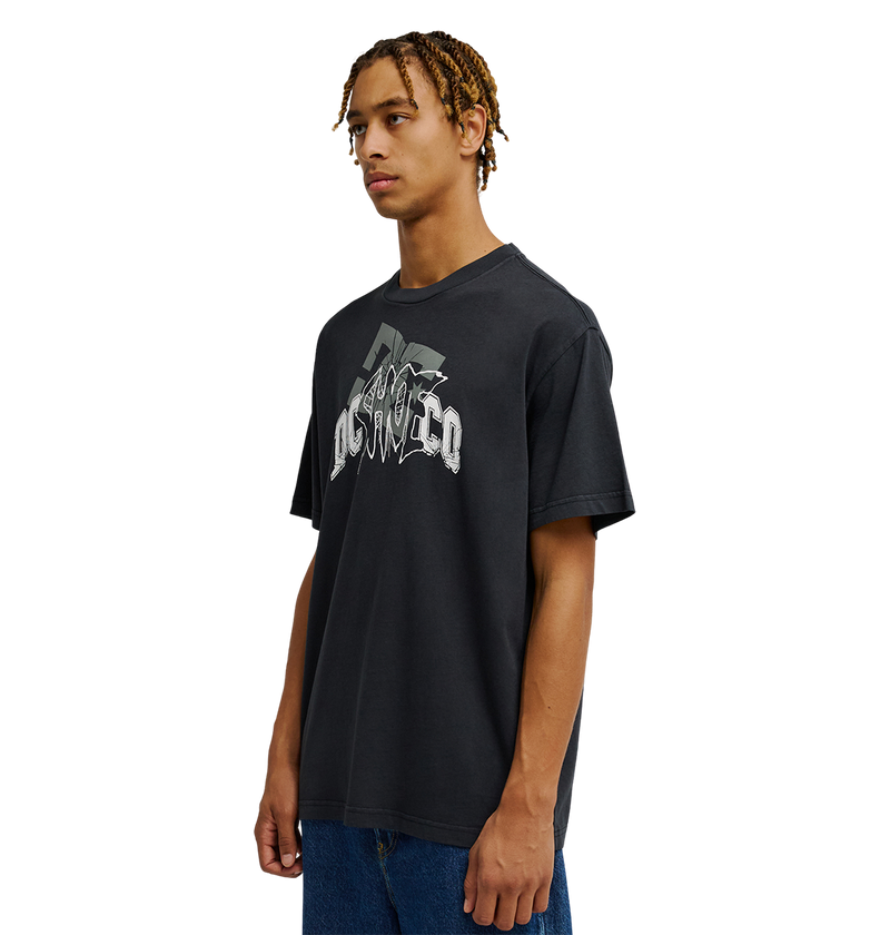 A young man with braided hair wears the Men's DC Speratic Tee by DC Shoes—a relaxed fit black graphic skate t-shirt featuring heritage-inspired artwork and stylized text—paired with blue jeans, standing against a plain white background.
