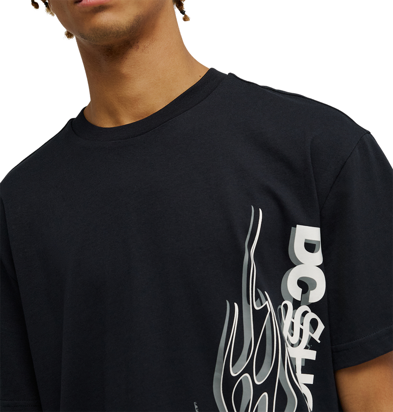 A person wearing the Men's DC Side Flame Tee by DC Shoes—a black graphic tee with a bold white stylized flame design and "DC SHOE" text on the front—shown from shoulders to mid-chest, perfect for skate sessions or showing off striking DC artwork.