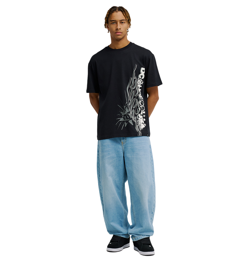 A person stands facing forward, wearing the Men's DC Side Flame Tee by DC Shoes—a black graphic tee with white abstract text and DC artwork-inspired designs—paired with loose light blue jeans and black sneakers, against a plain white background.