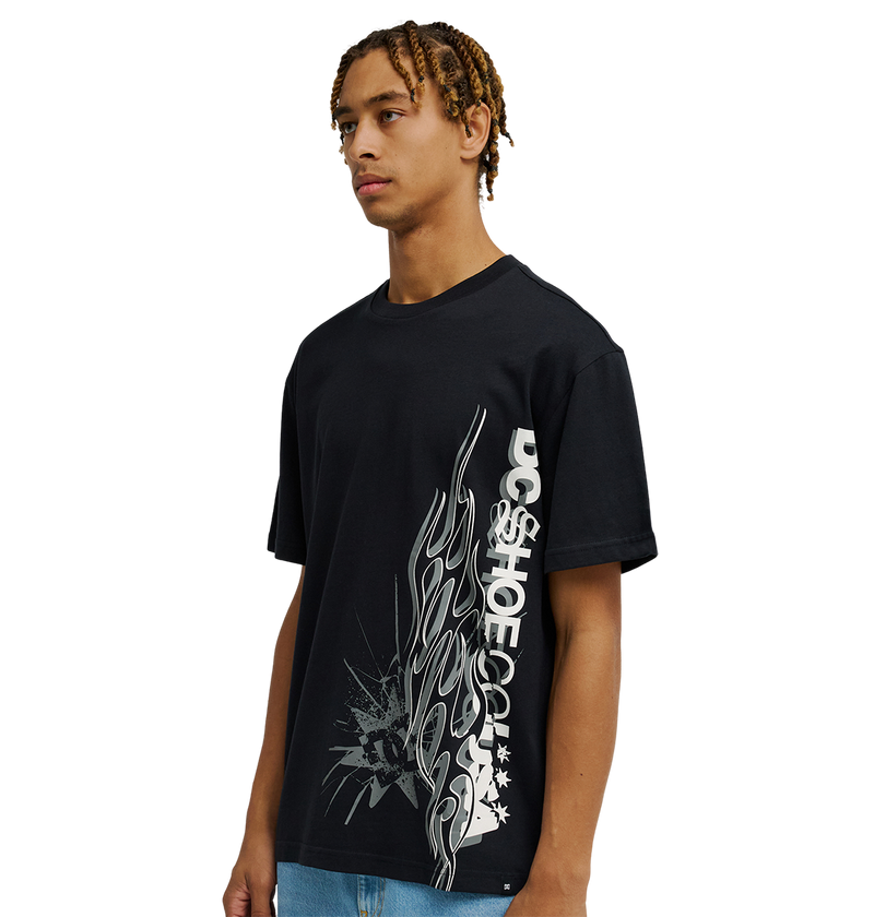 A young man stands against a plain white background wearing the Men's DC Side Flame Tee by DC Shoes, a black t-shirt featuring bold white flame graphics and text on the front—an ideal choice for skate sessions.