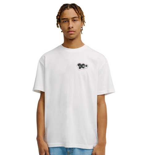 A person with short, light brown braids wears the Men's DC Side Flame Tee from DC Shoes, a relaxed fit white skate shirt featuring a small black and white graphic on the left chest, standing against a plain white background.