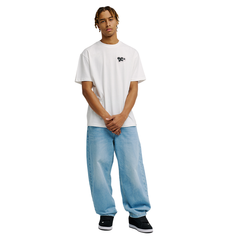 A person with short dreadlocks wears the Men's DC Side Flame Tee by DC Shoes—a relaxed fit white graphic skate tee—paired with light blue baggy jeans and black sneakers, standing against a plain white background with hands loosely clasped in front.