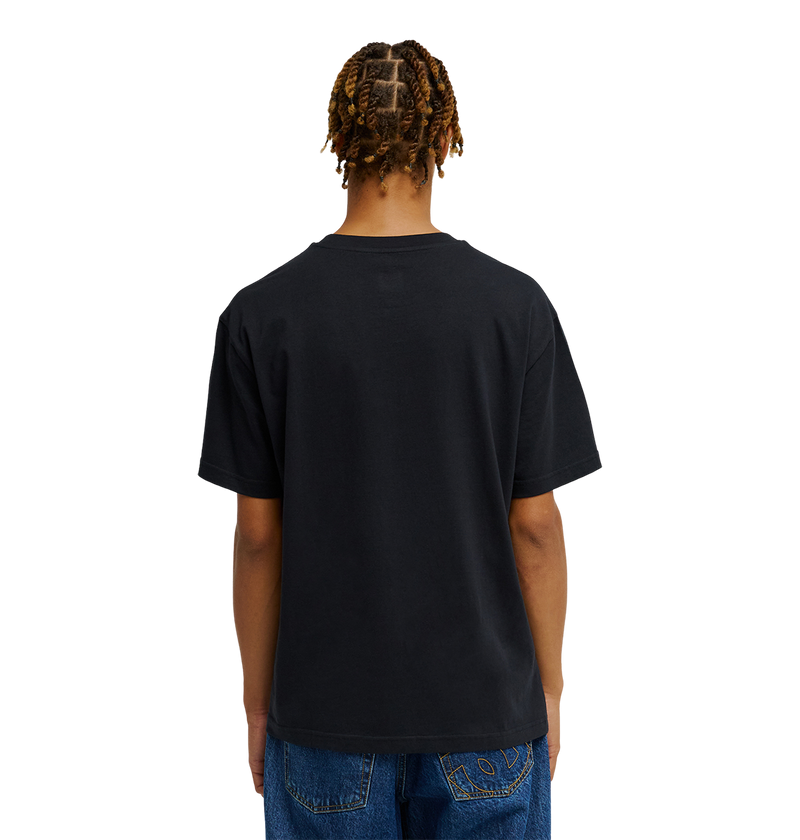 A person with braided hair is shown from the back against a plain white background, wearing blue jeans and the Men's DC Heritage Font Tee by DC Shoes in a black relaxed fit graphic skate style.