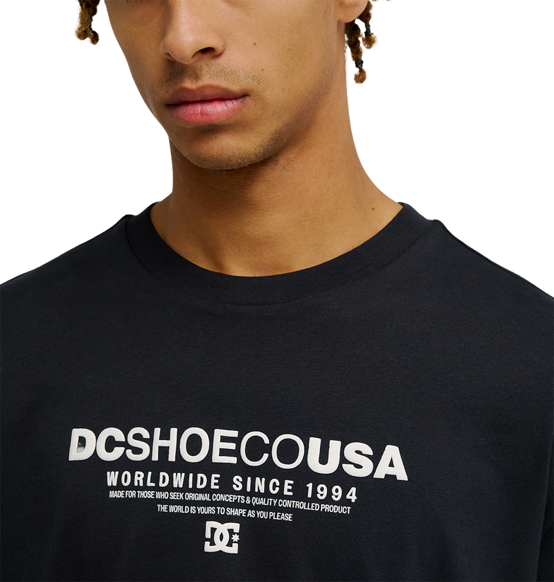 A person wearing the Men's DC Heritage Font Tee by DC Shoes, featuring the bold "DCSHOECOUSA Worldwide Since 1994" graphic across the chest. The photo shows only the lower half of the face and upper torso, emphasizing the relaxed fit of this black skate t-shirt.