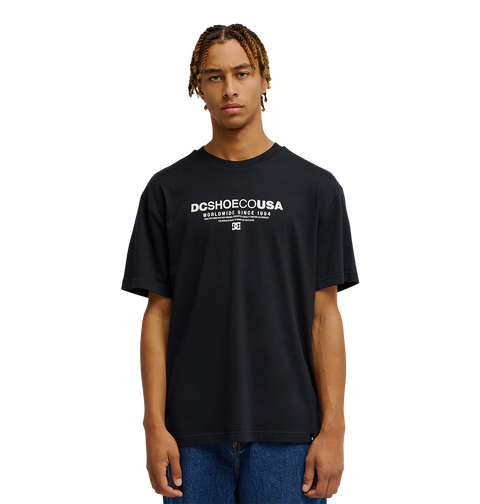 A person with short, braided hair wears the Men's DC Heritage Font Tee by DC Shoes in black, styled in a relaxed fit with blue jeans, standing against a plain white background.
