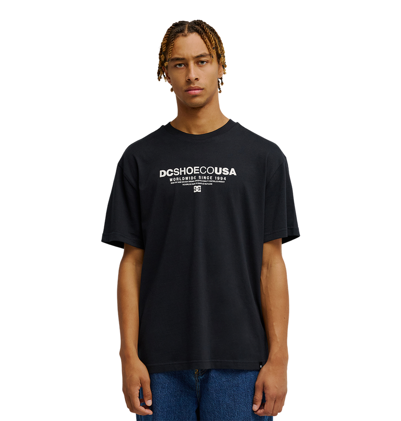 A person with short, braided hair wears the Men's DC Heritage Font Tee by DC Shoes in black, styled in a relaxed fit with blue jeans, standing against a plain white background.