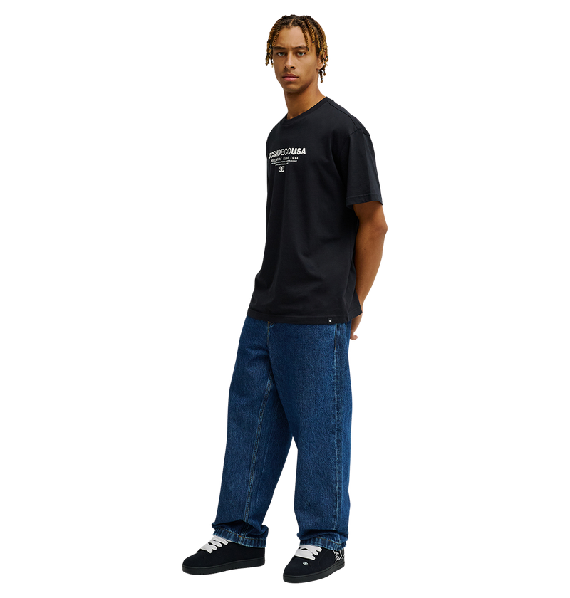 A person stands against a white background wearing the Men's DC Heritage Font Tee by DC Shoes, styled in black with a bold heritage font graphic. They pair it with relaxed-fit loose blue jeans and black sneakers. With short braided hair, they look slightly to the side, hands tucked in their pockets.