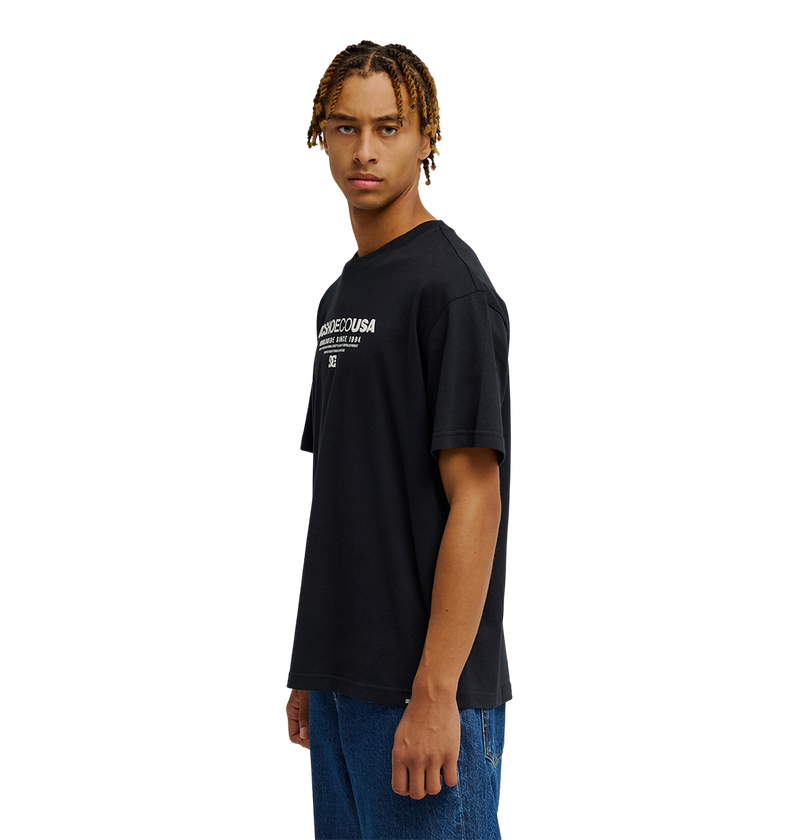 A person with short, braided hair wears the Men's DC Heritage Font Tee by DC Shoes—a relaxed fit graphic skate t-shirt with white heritage font—paired with blue jeans, standing sideways and looking forward against a plain white background.