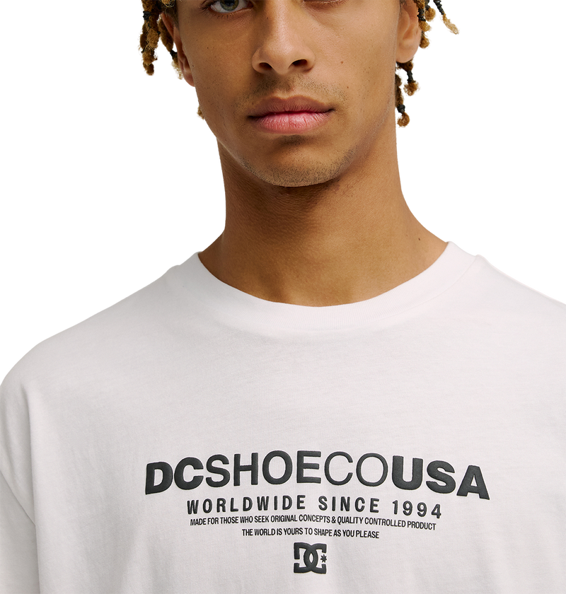 A person wearing the Men's DC Heritage Font Tee by DC Shoes, a white graphic skate t-shirt featuring bold black text that reads "WORLDWIDE SINCE 1994" with additional smaller lettering beneath. The photo shows only the lower face and upper torso, drawing attention to the tee's relaxed fit and classic skate style.
