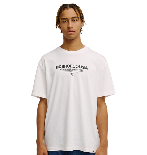 A young man models the Men's DC Heritage Font Tee by DC Shoes—a relaxed fit white graphic skate shirt featuring "DCSHOECOUSA Worldwide Since 1994" printed in a heritage font on the front—as he stands against a plain white background, gazing straight ahead with a neutral expression.