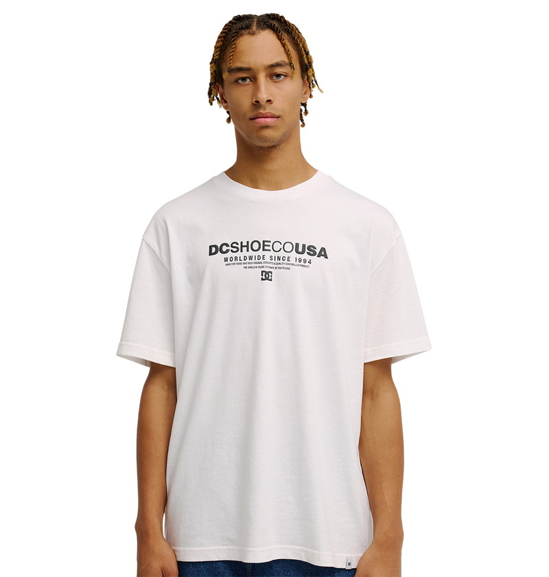 A young man models the Men's DC Heritage Font Tee by DC Shoes—a relaxed fit white graphic skate shirt featuring "DCSHOECOUSA Worldwide Since 1994" printed in a heritage font on the front—as he stands against a plain white background, gazing straight ahead with a neutral expression.