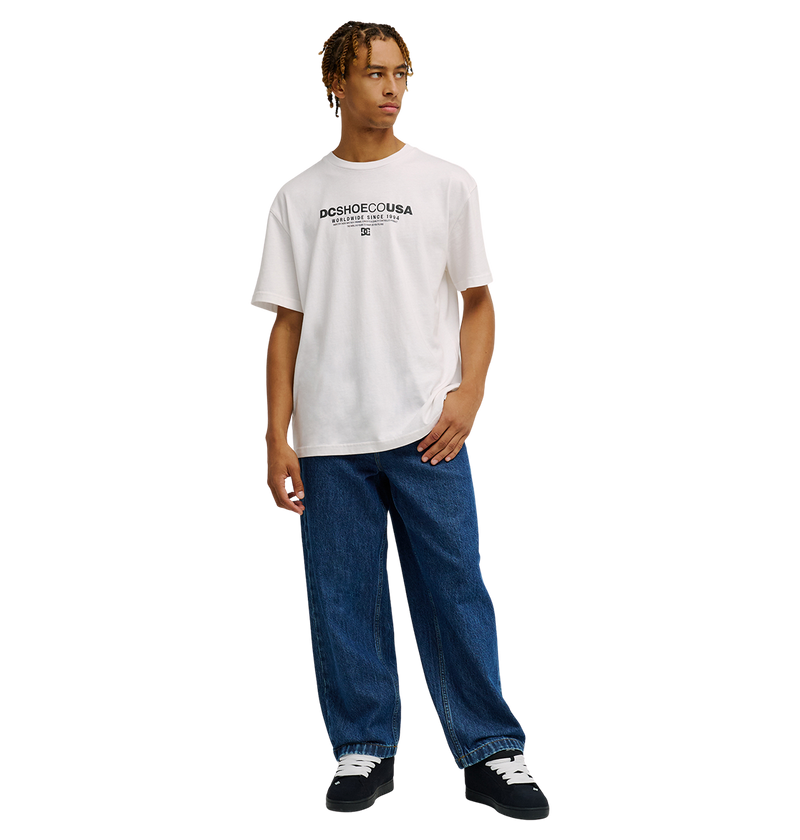 A person stands against a plain white background, wearing the Men's DC Heritage Font Tee by DC Shoes in a relaxed fit with a white graphic design, paired with loose blue jeans and black sneakers with white soles.
