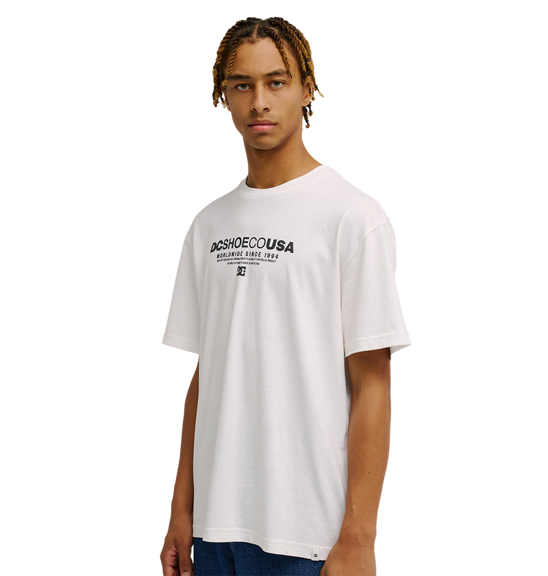 A young person with braided hair wears the Men's DC Heritage Font Tee by DC Shoes—a relaxed fit white graphic skate shirt—paired with blue jeans, standing against a plain white background.